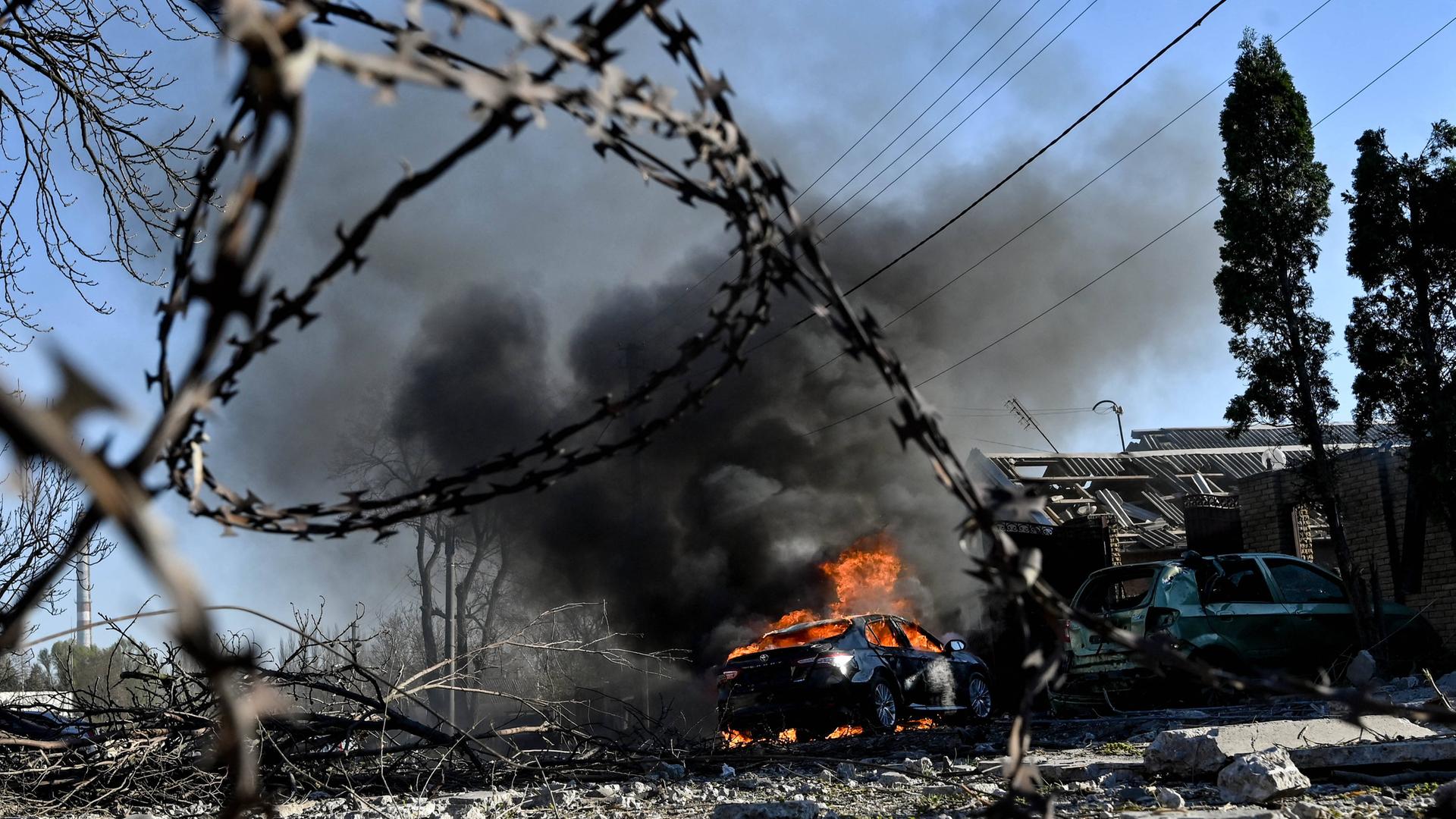 Das Bild aus der ukrainischen Region Saporischschja zeigt zerstörte Gebäude und ein brennendes Auto, Rauch steigt auf. Im Vordergrund ist Stacheldraht zu sehen. 