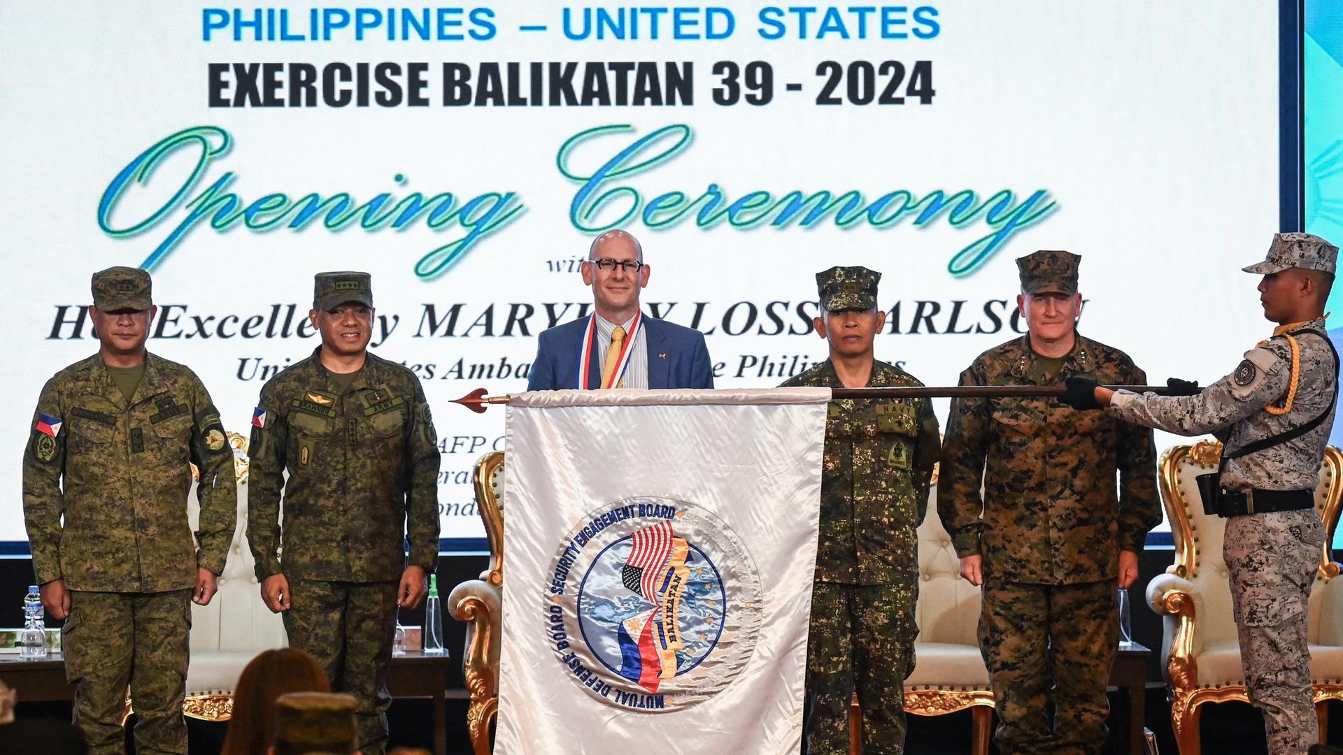 Philippinische und amerikanische Militärs stehen bei der Eröffnungszeremonie des Militärmanövers Exercise Balikatan 39 - 2024 nebeneinander. Ein Soldat hält eine Fahne mit den Flaggen beider Länder waagerecht vor den Militärs.