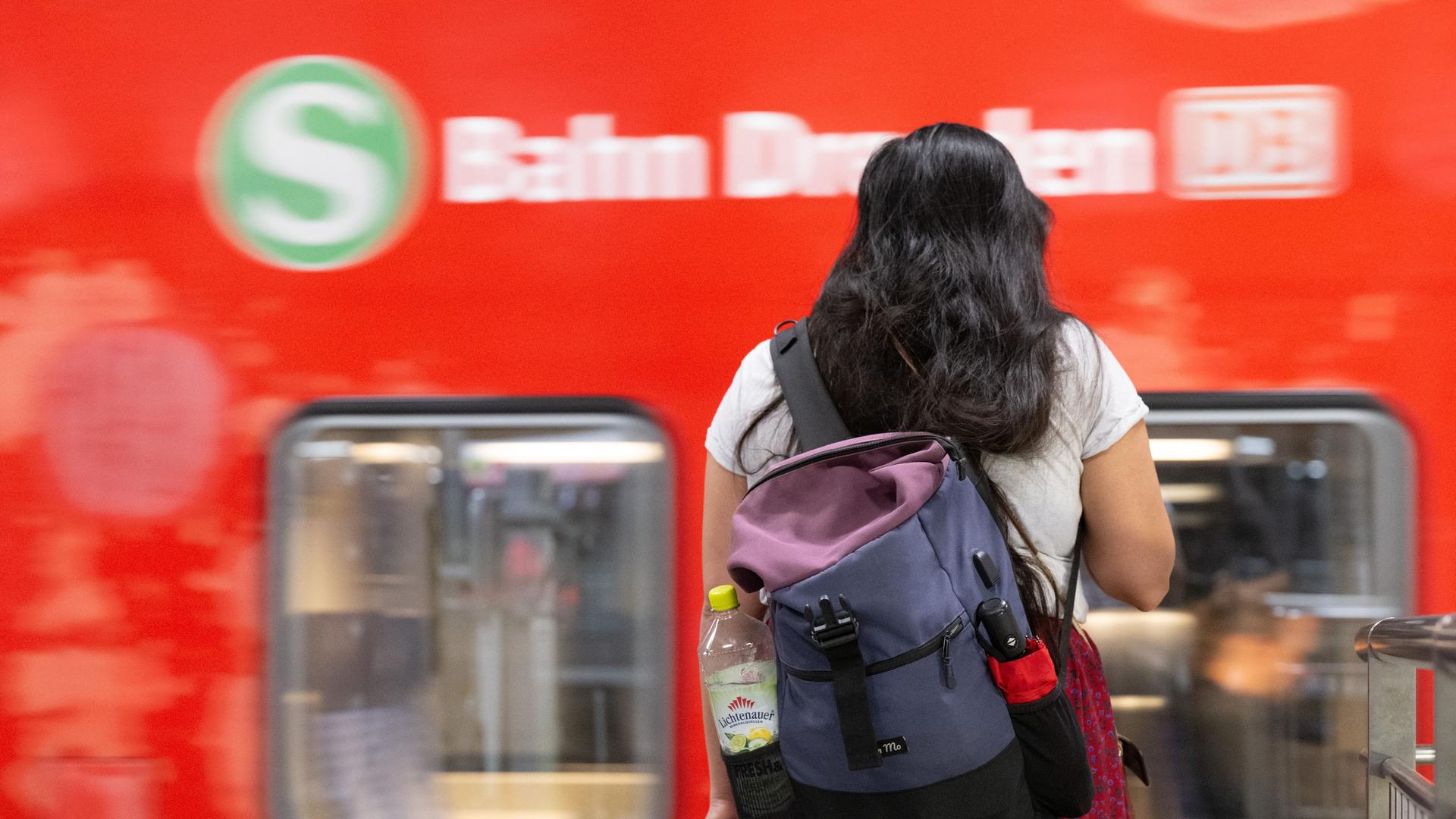 Eine Frau steht im Hauptbahnhof auf einem Bahnsteig vor einer S-Bahn. Sie dreht der Kamera den Rücken zu und trägt einen Rucksack.
