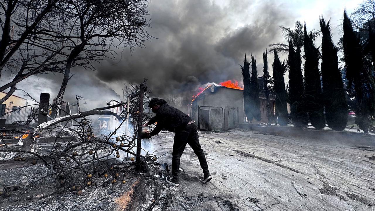 Ein Mann versucht, einen Brand zu löschen, während um ihn herum verkohlte Baumreste und im Hintergrund ein brennendes Haus zu sehen sind.