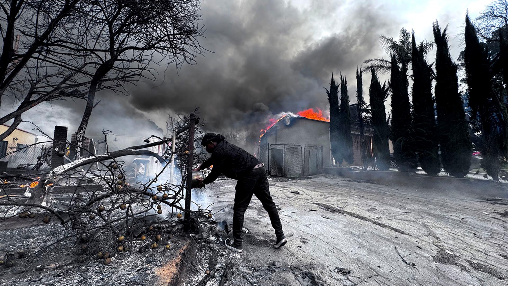 Ein Mann versucht, einen Brand zu löschen, während um ihn herum verkohlte Baumreste und im Hintergrund ein brennendes Haus zu sehen sind.