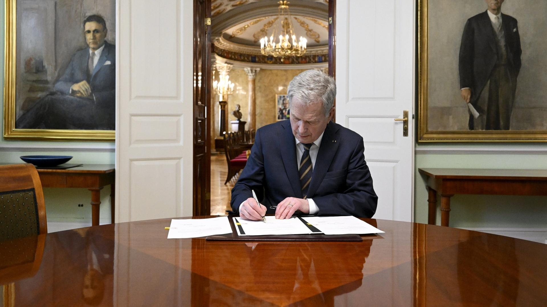 Niinistö sitzt an einem braunen Tisch und unterzeichnet gerade die Dokumente. Hinter ihm eine offene Flügeltür und Wände mit zwei großen Portraits seiner Vorgänger. Niinistö sitzt an einem braunen Tisch und unterzeichnet gerade die Dokumente. Hinter ihm eine offene Flügeltür und Wände mit zwei großen Portraits seiner Vorgänger.