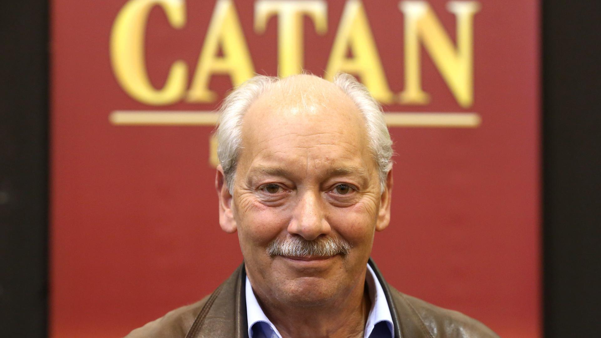 Klaus Teuber, Erfinder des Strategiespiels "Die Siedler von Catan", trägt Lederjacke und Hemd, Haarkranz und Schnäuzer. hinter hm steht auf rotem Grund "Catan". Klaus Teuber, Erfinder des Strategiespiels "Die Siedler von Catan", trägt Lederjacke und Hemd, Haarkranz und Schnäuzer. hinter hm steht auf rotem Grund "Catan".