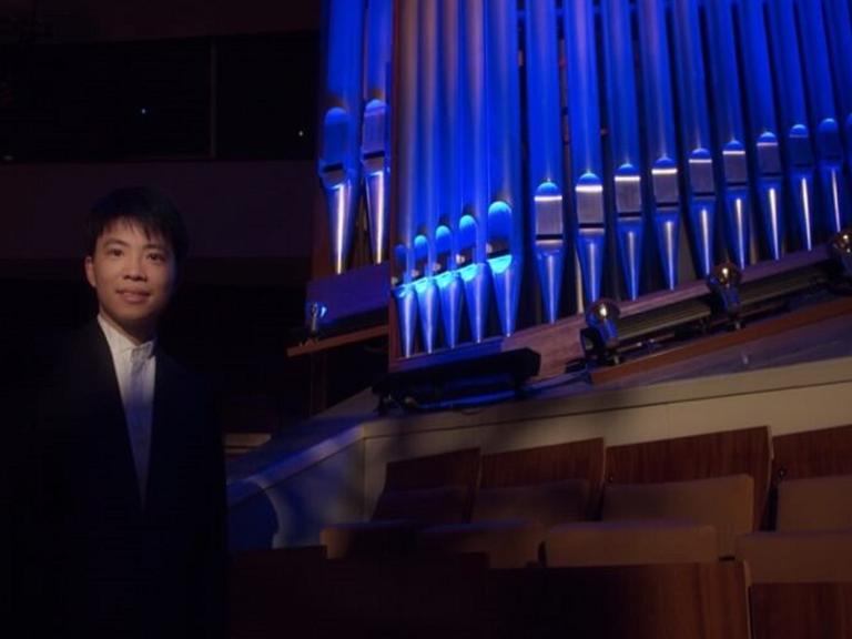 Kit Armstrong, ein junger Mann, steht neben einer blau beleuchteten Orgel und lächelt in die Kamera.