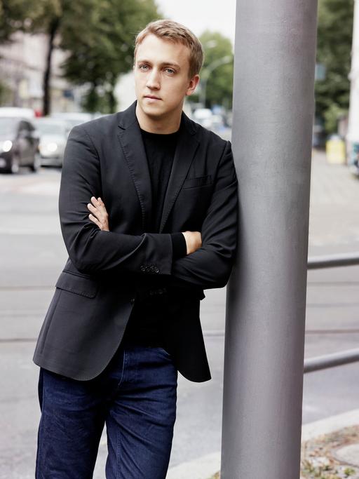 Ein junger Mann mit blondem Haar lehnt mit verschränkten Armen an einem silbergrauen Laternenpfahl. Er trägt Jeans und ein schwarzes Jackett. Im Hintergrund ist eine Straße mit geparkten Autos und Bäumen zu sehen.