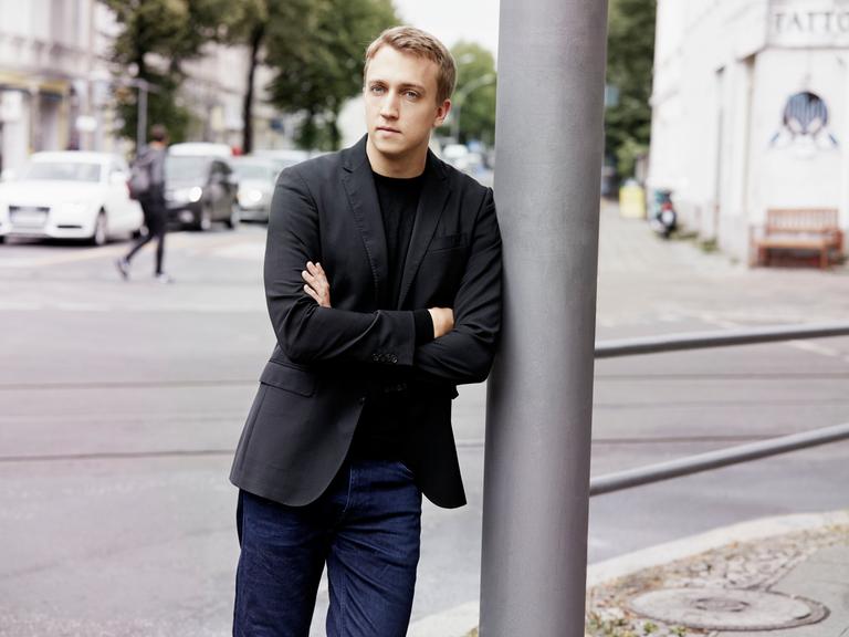 Ein junger Mann mit blondem Haar lehnt mit verschränkten Armen an einem silbergrauen Laternenpfahl. Er trägt Jeans und ein schwarzes Jackett. Im Hintergrund ist eine Straße mit geparkten Autos und Bäumen zu sehen.