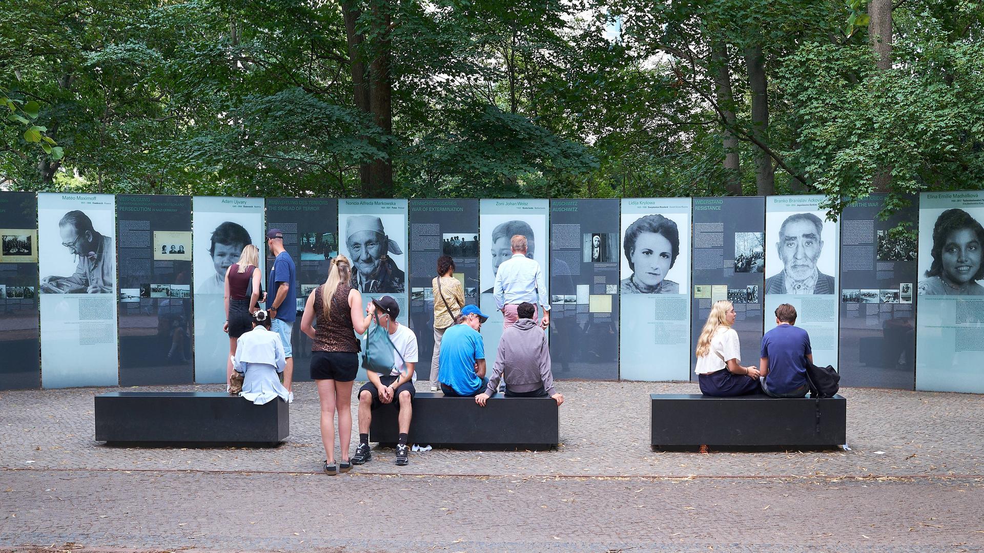 Menschen sitzen auf Bänken vor einer Reihe von Fotos und Tafeln, die an ermordete Sinti und Roma erinnern. Menschen sitzen auf Bänken vor einer Reihe von Fotos und Tafeln, die an ermordete Sinti und Roma erinnern.