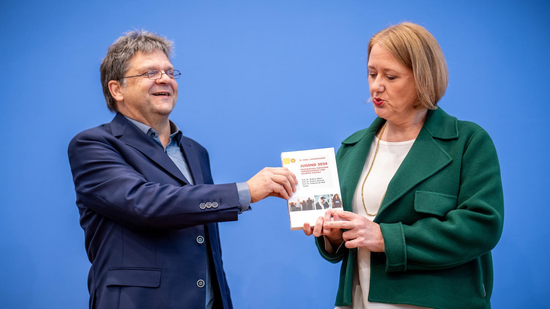 Links im Bild steht ein Mann im dunkelblauem Anzug und hält seine Hand an ein Buch. Dieses wird von einer Frau, Lisa Paus, die ein grünes Jacket und ein weißes T-Shirt trägt, in ihren Händen gehalten. Sie stehen vor einer blauen Wand.