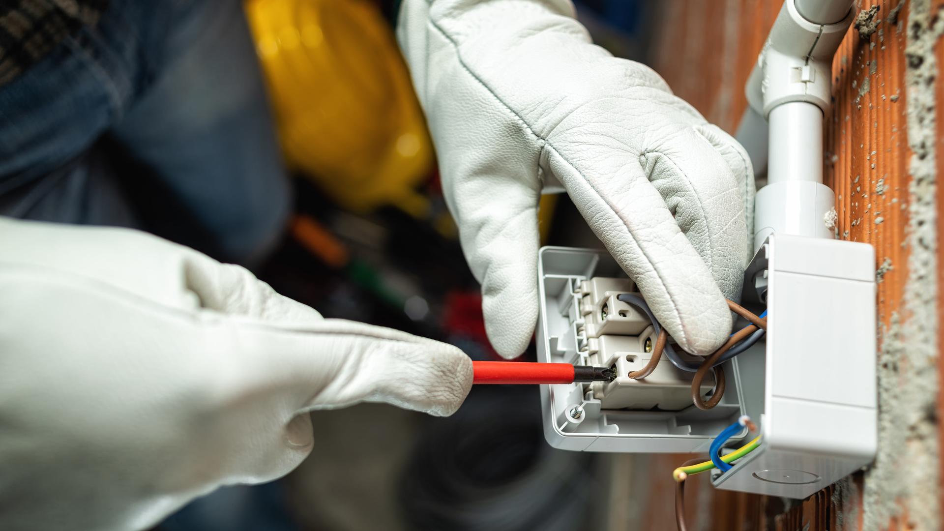 Ein Elektriker schraubt an einem Schalter, zu sehen sind nur die Hände in Handschuhen.