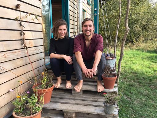 Eine junge Frau und ein junger Mann sitzen auf gestapelten Europaletten, die den Eingang zu ihrem Tiny House bilden, dessen längere Seite links von ihnen zu sehen ist. An den Rändern des Aufgangs stehen Blumentöpfe mit Blumen. Beide sind barfuß, die Frau trägt schwarze Kleidung, der Mann eine grau Hose und ein weinrotes Oberteil. Beide strahlen.