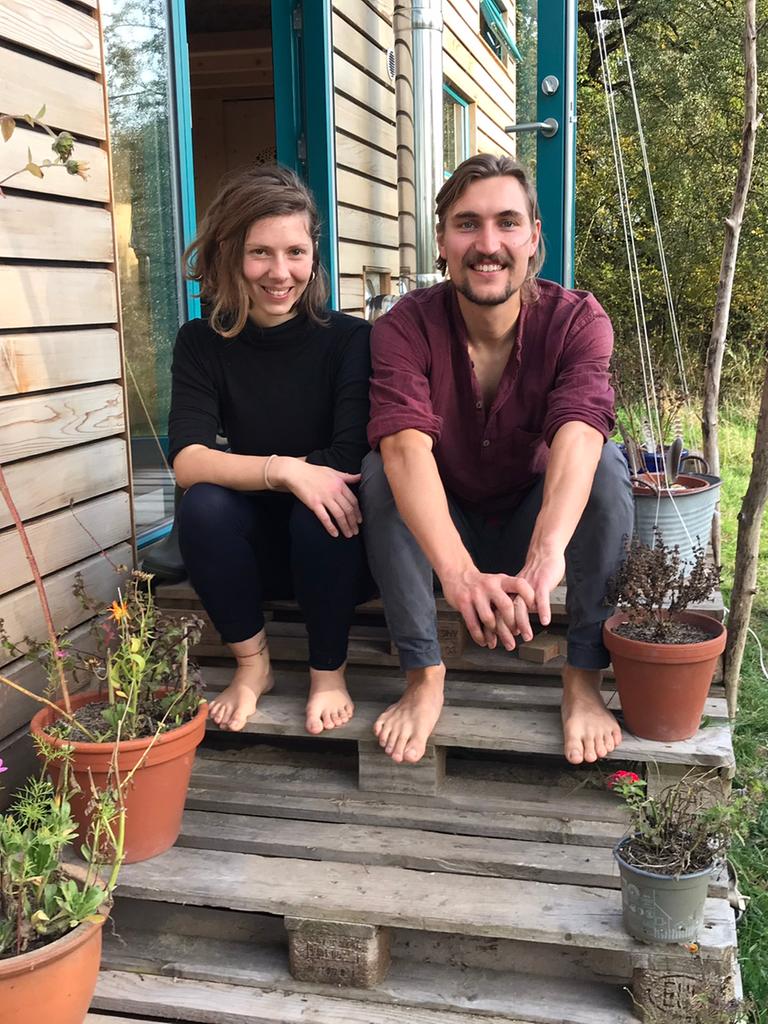Eine junge Frau und ein junger Mann sitzen auf gestapelten Europaletten, die den Eingang zu ihrem Tiny House bilden, dessen längere Seite links von ihnen zu sehen ist. An den Rändern des Aufgangs stehen Blumentöpfe mit Blumen. Beide sind barfuß, die Frau trägt schwarze Kleidung, der Mann eine grau Hose und ein weinrotes Oberteil. Beide strahlen.