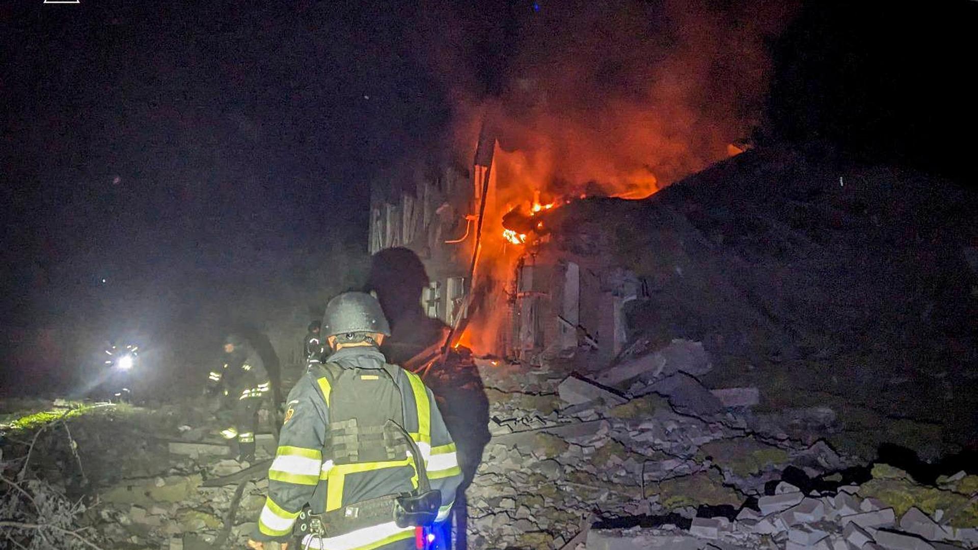 Es ist dunkel. Vor einem brennenden Gebäude sieht man einen angeleuchteten Feuerwehrmann, dessen Schatten auf Trümmer fällt. Es ist dunkel. Vor einem brennenden Gebäude sieht man einen angeleuchteten Feuerwehrmann, dessen Schatten auf Trümmer fällt.