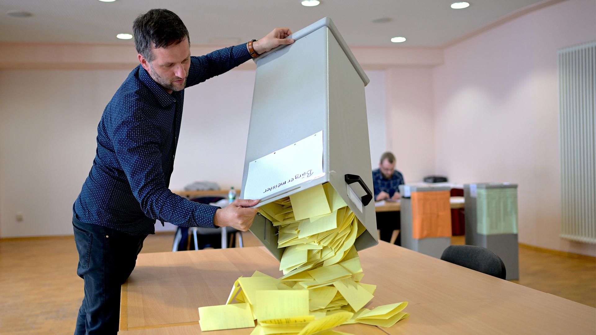 Aus einer großen Kiste schüttet ein Mann die ausgefüllten Wahl-Zettel auf einen Tisch.