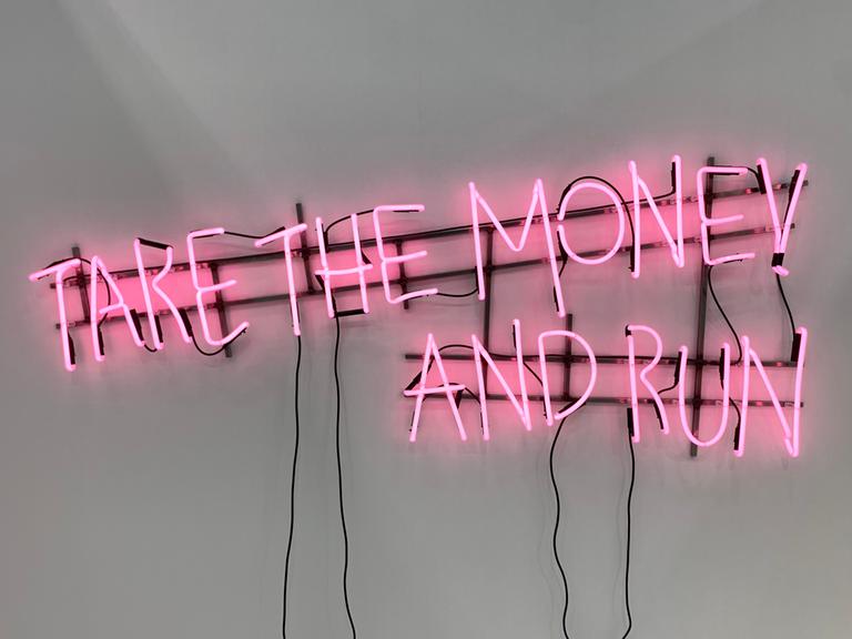 Das Werk „Ne travallez jemais“ (2022) von Kerim Seiler am Stand der Galerie Sexauer auf der Art Cologne. In rosa Buchstaben steht in mit Leuchtstoffröhren vor weisser Wand geschrieben "Take the money and run".