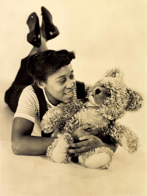 Historisches Sepiafoto von Leila Negra, die mit einem Teddybären auf dem Boden liegt.
