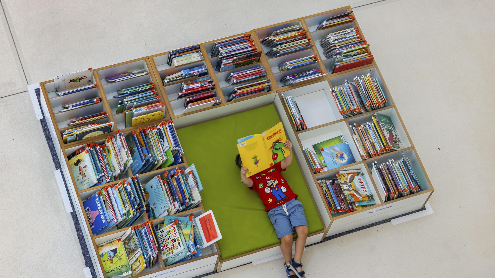Ein Kind liegt auf dem Boden mit einem Buch in der Hand umgeben von Buchregal mit Büchern. Ein Kind liegt auf dem Boden mit einem Buch in der Hand umgeben von Buchregal mit Büchern.