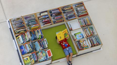 Ein Kind liegt auf dem Boden mit einem Buch in der Hand umgeben von Buchregal mit Büchern.