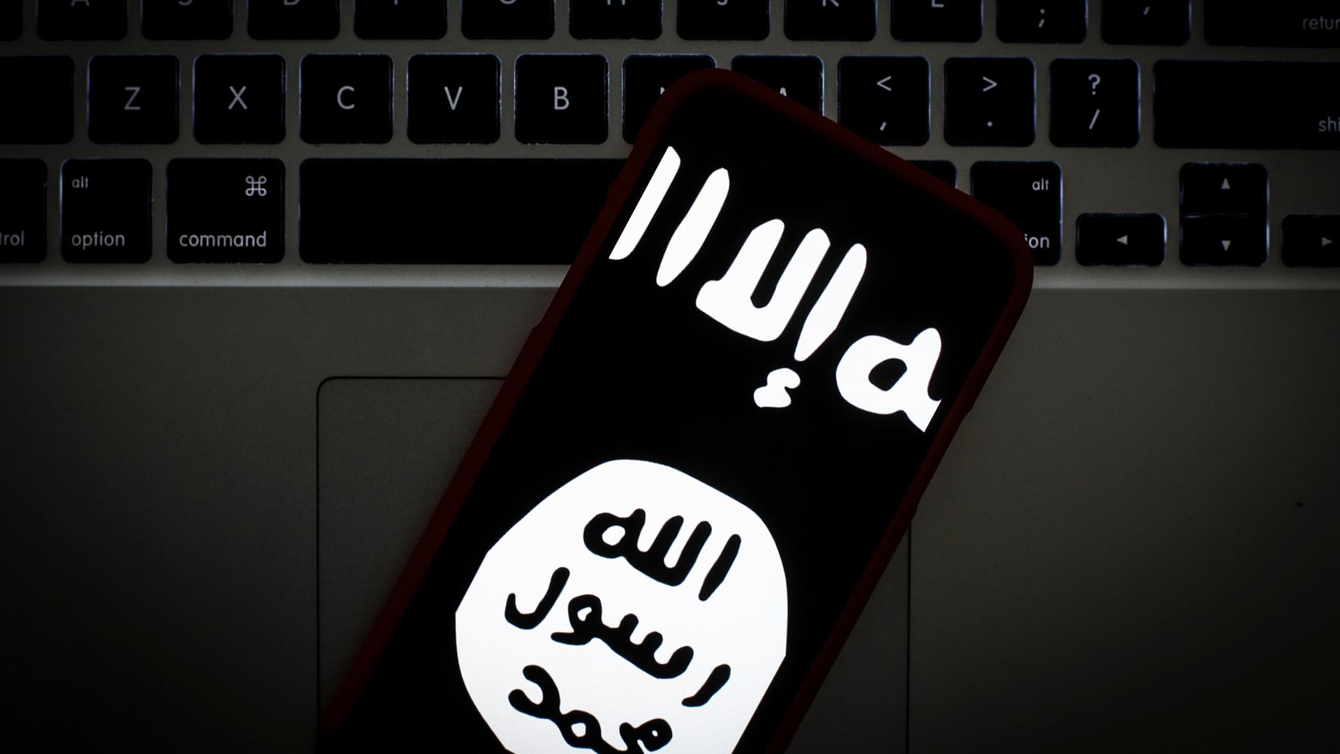 Das Logo des islamischen Staates auf dem Bildschirm eines Smartphones. Im Hintergrund ein Laptop.