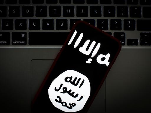 Das Logo des islamischen Staates auf dem Bildschirm eines Smartphones. Im Hintergrund ein Laptop.