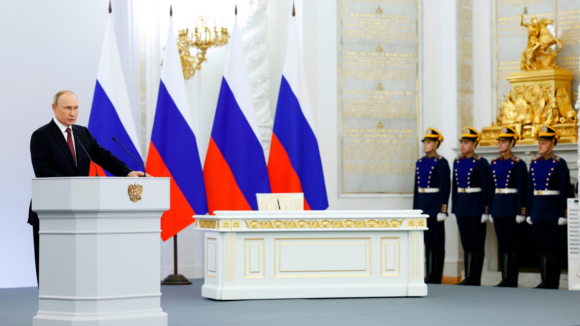 Wladimir Putin steht an einem Rednerpult. Hinter ihm vier russische Flaggen. Neben ihm stehen vier Soldaten.