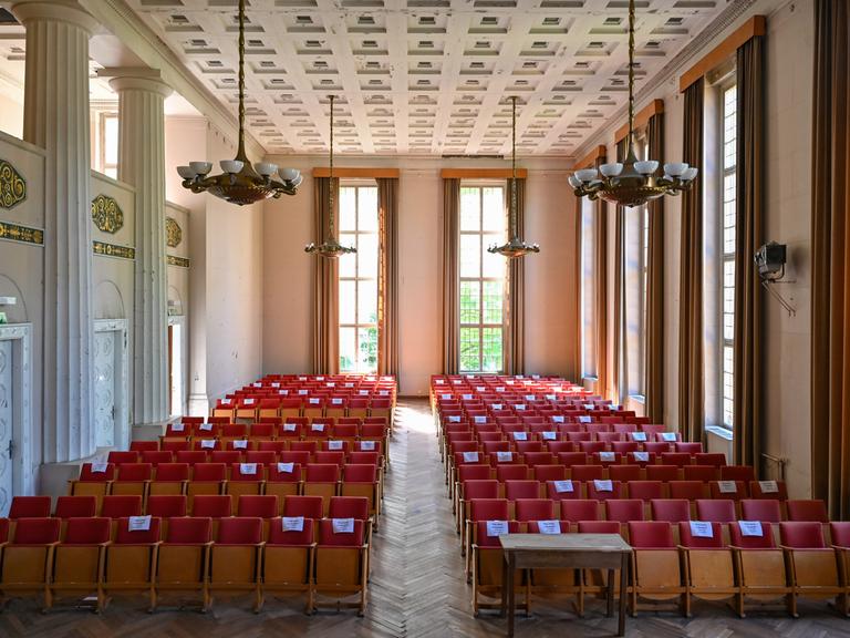 Ein großer Saal  mit hoher Decke und hohen Fenstern. Von der Decke hängen große Leuchter. Das Parkett ist mit roten Stühlen ausgestattet.