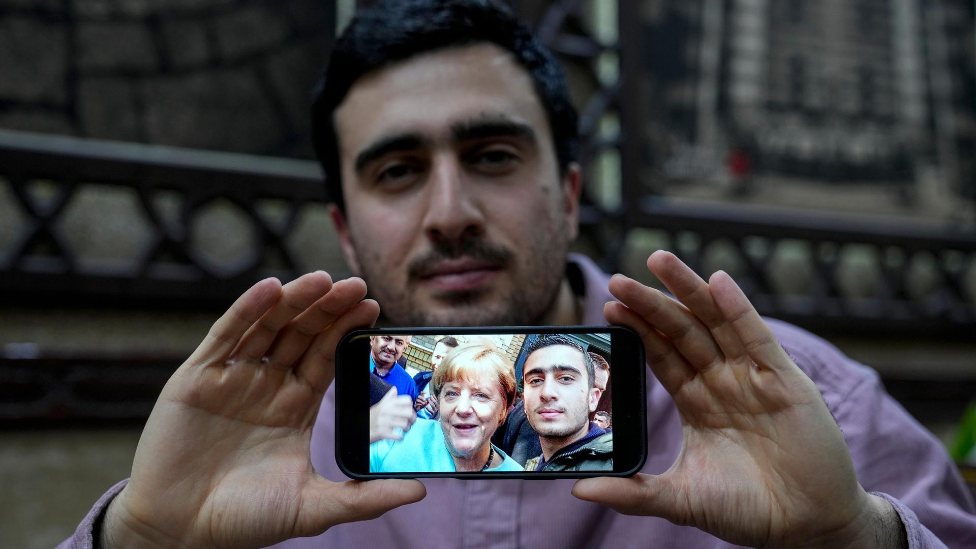 Anas Modamani hält ein Handy mit seinem Selfie mit Angela Merkel mit zwei Händen vor seinem Körper.