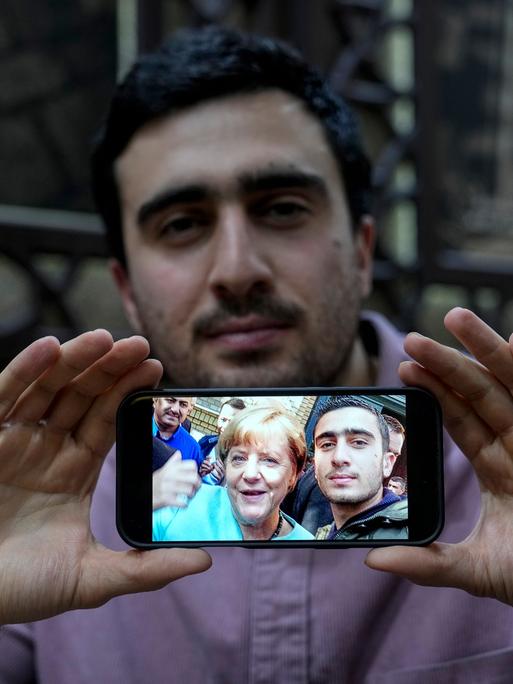 Anas Modamani hält ein Handy mit seinem Selfie mit Angela Merkel mit zwei Händen vor seinem Körper.