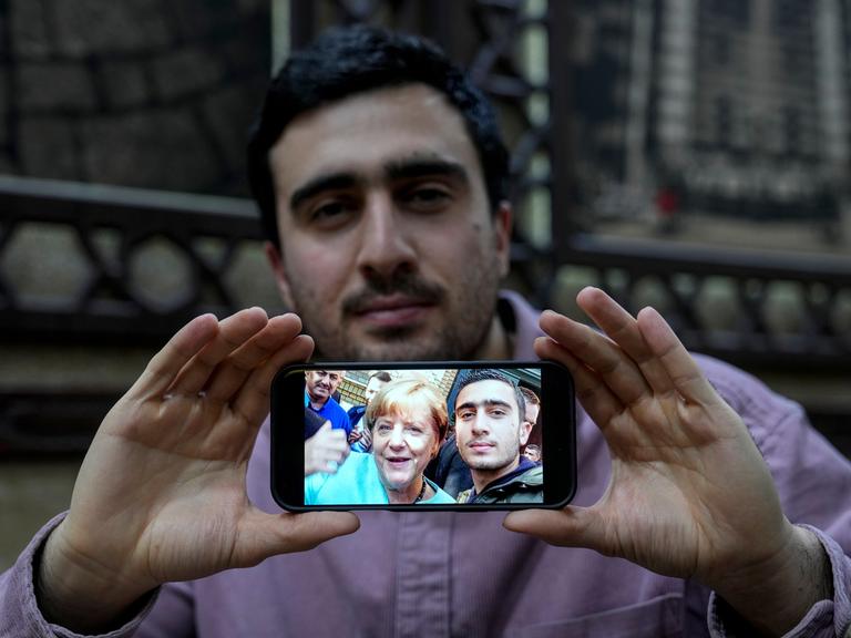 Anas Modamani hält ein Handy mit seinem Selfie mit Angela Merkel mit zwei Händen vor seinem Körper.
