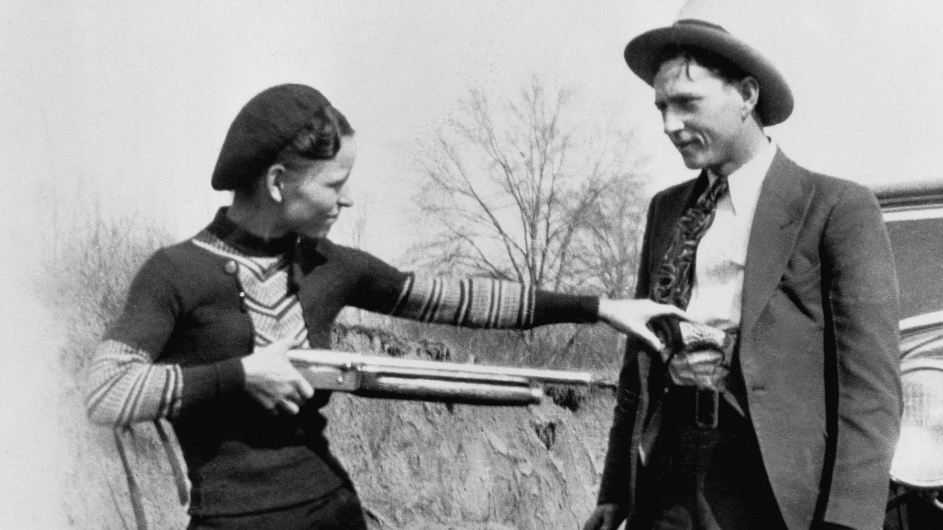 Bonnie Parker in dunklem Kleid und Clyde Barrow im Anzug, posieren mit einer Flinte vor einem Auto.