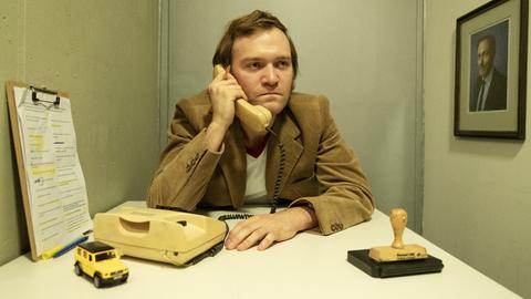 Ein Mann im 70er-Jahre Anzug sitzt an einem Tisch und hält einen Telefonhörer in der Hand. Er sieht nicht sehr glücklich aus. Vor ihm liegt ein Stempelkissen und ein Spielzeugjeep.