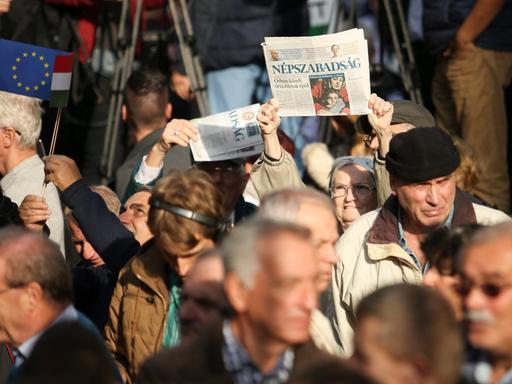 Inmitten einer Gruppe Demonstrierender Menschen, hält eine ältere Frau eine Print-Ausgabe der Zeitung "Nepszabadsag" in die Luft.