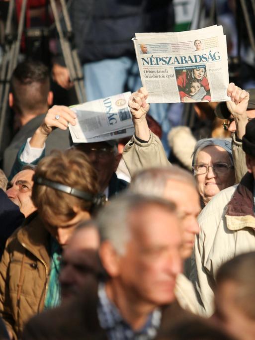 Inmitten einer Gruppe Demonstrierender Menschen, hält eine ältere Frau eine Print-Ausgabe der Zeitung "Nepszabadsag" in die Luft.