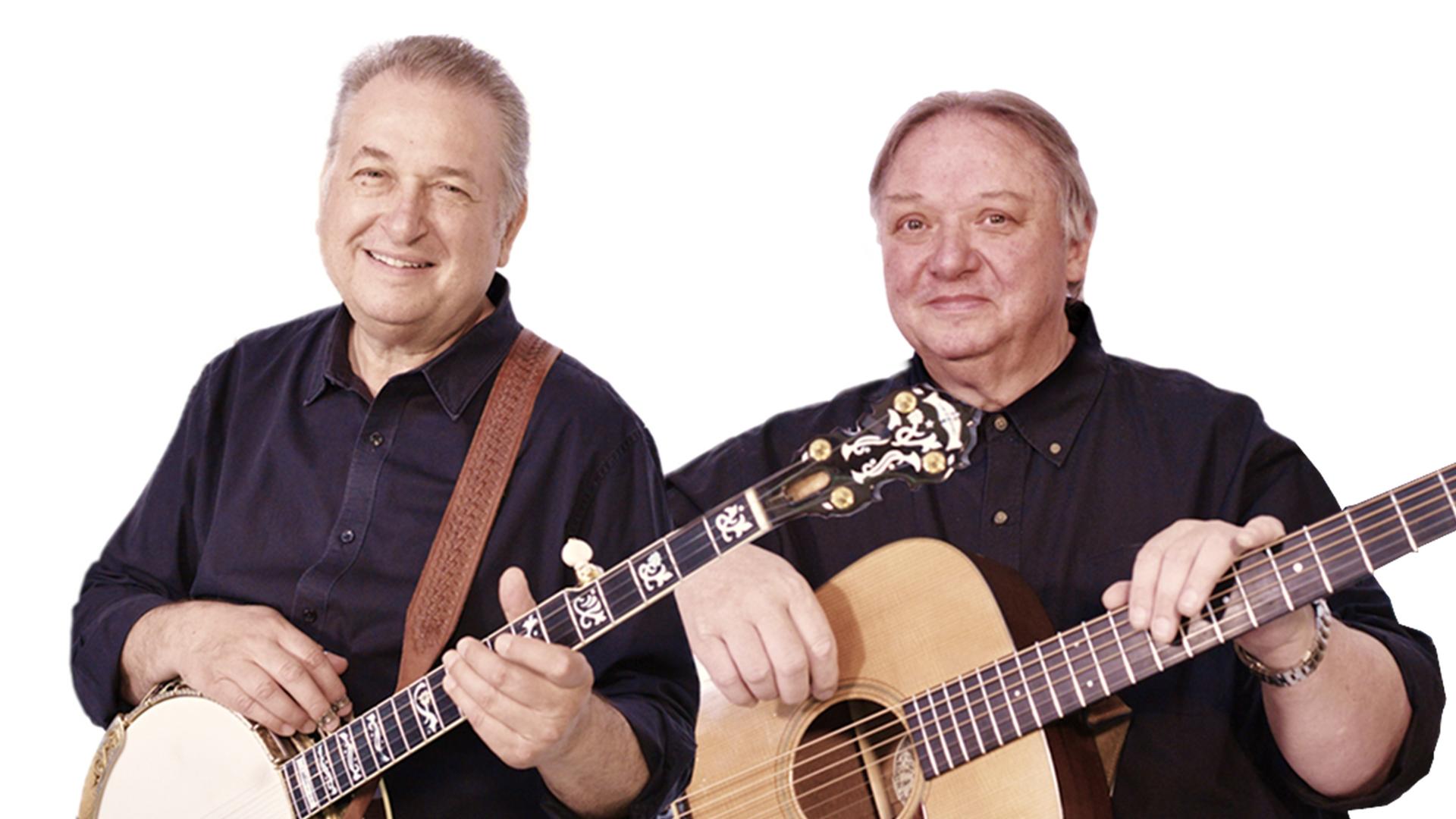 Zwei Männer mit kurzen grauen Haaren posieren mit Banjo (links) und Gitarre (rechts) für die Kamera. Sie tragen dunkle Hemden und lächeln.