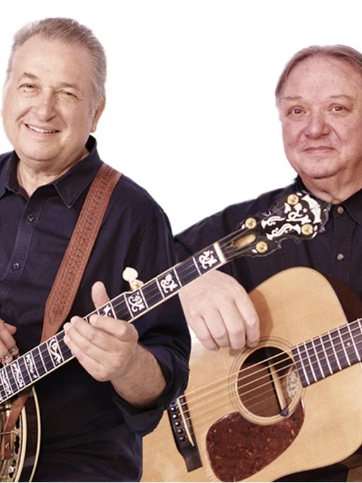 Zwei Männer mit kurzen grauen Haaren posieren mit Banjo (links) und Gitarre (rechts) für die Kamera. Sie tragen dunkle Hemden und lächeln.