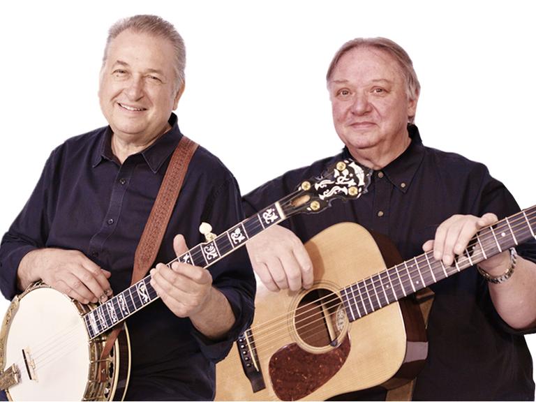 Zwei Männer mit kurzen grauen Haaren posieren mit Banjo (links) und Gitarre (rechts) für die Kamera. Sie tragen dunkle Hemden und lächeln.