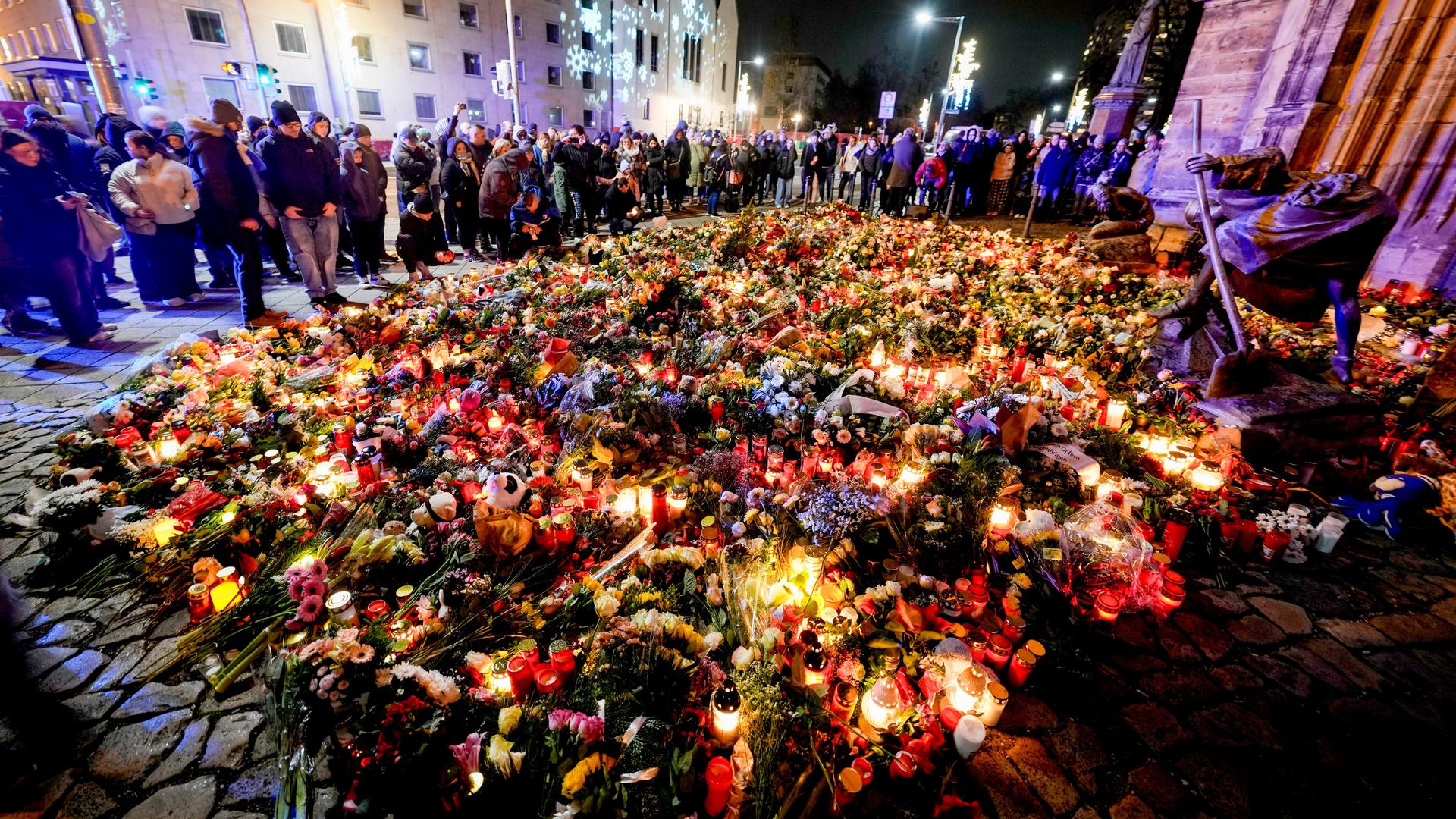 Trauernde Menschen stehen um eine große Fläche von Blumen und Kerzen herum vor dem Portal der Johanniskirche