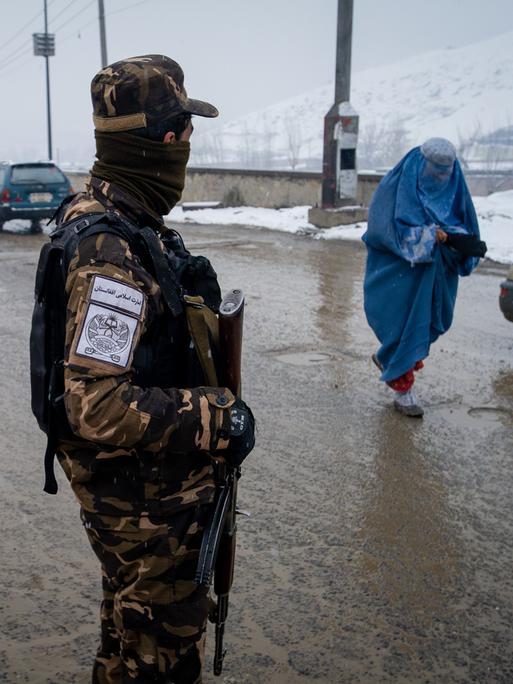 Eine afghanische Frau in blauer Burka bettelt im Schneegestäuber an einem Kontrollpunkt der Straße von Kabul nach Pul-e Alam, beobachtet von einem bewaffneten Taliban.