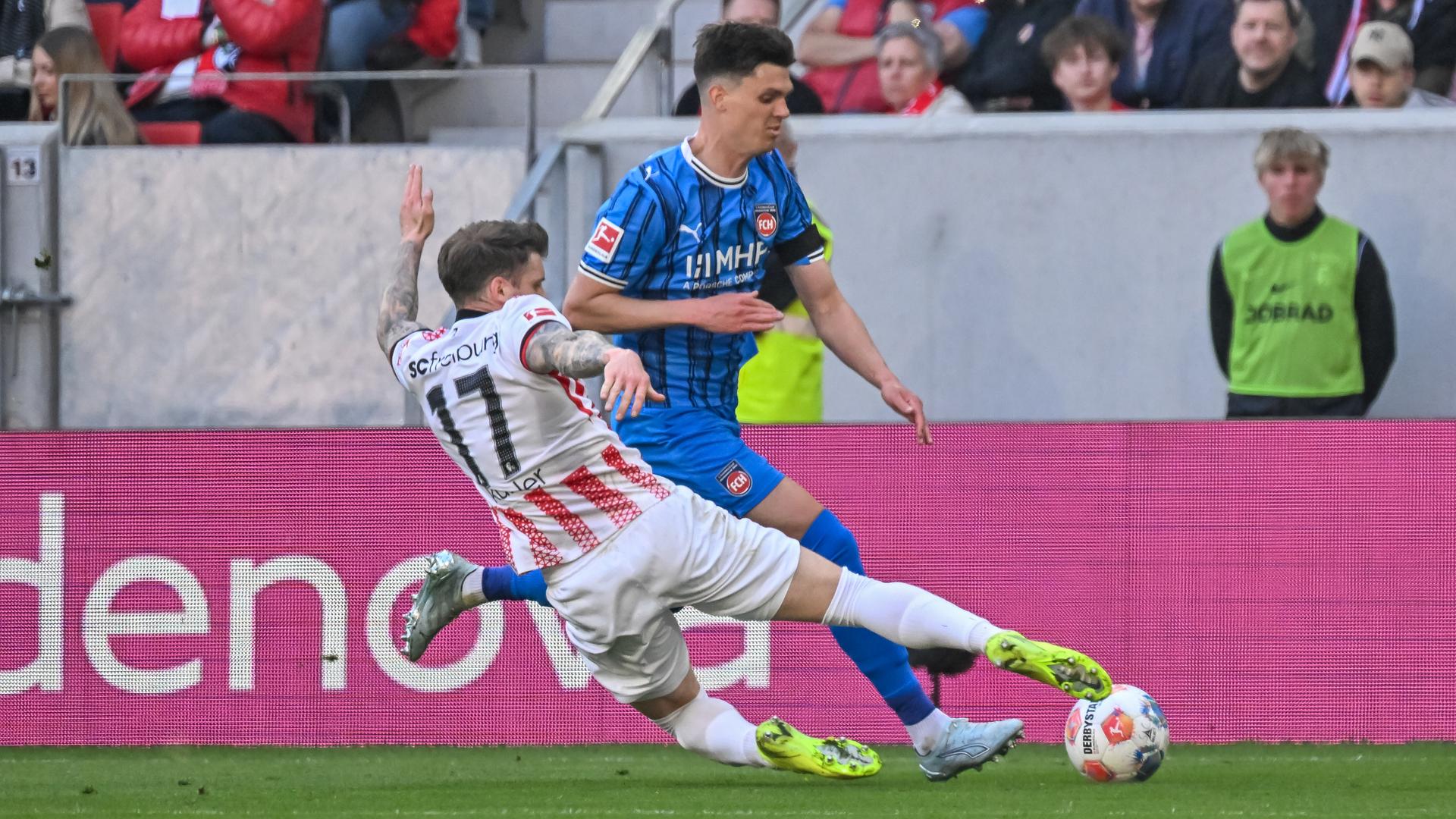 Kübler im rot-weißen Trikot sieht man von hinten. Er grätscht nach dem Ball, den Honsak im blau-schwarzen Trikot an ihm vorbeispielen will. Dahinter eine Zuschauertribüne.