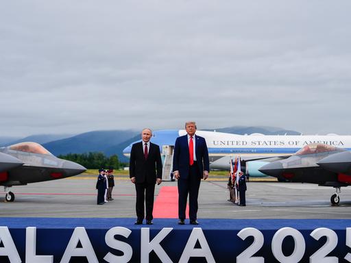 US-Präsident Donald Trump und der russische Präsident Wladimir Putin stehen auf einem Flughafen auf einem Podest, auf dem "Alaska 2025" steht.