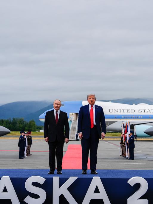 US-Präsident Donald Trump und der russische Präsident Wladimir Putin stehen auf einem Flughafen auf einem Podest, auf dem "Alaska 2025" steht.
