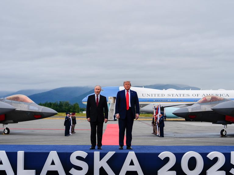 US-Präsident Donald Trump und der russische Präsident Wladimir Putin stehen auf einem Flughafen auf einem Podest, auf dem "Alaska 2025" steht.