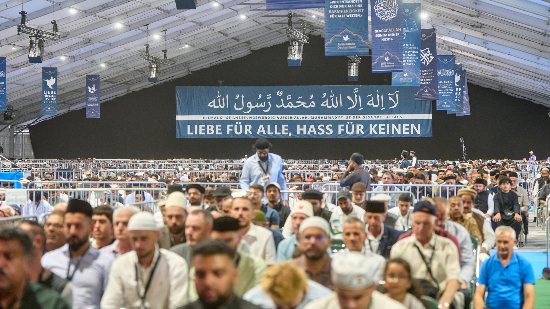 In einem großen Zelt sitzen muslimische Männer zum Gebet. Im Hintergrund ist ein großes Banner. Darauf steht: Liebe für alle, Hass für keinen. In einem großen Zelt sitzen muslimische Männer zum Gebet. Im Hintergrund ist ein großes Banner. Darauf steht: Liebe für alle, Hass für keinen.