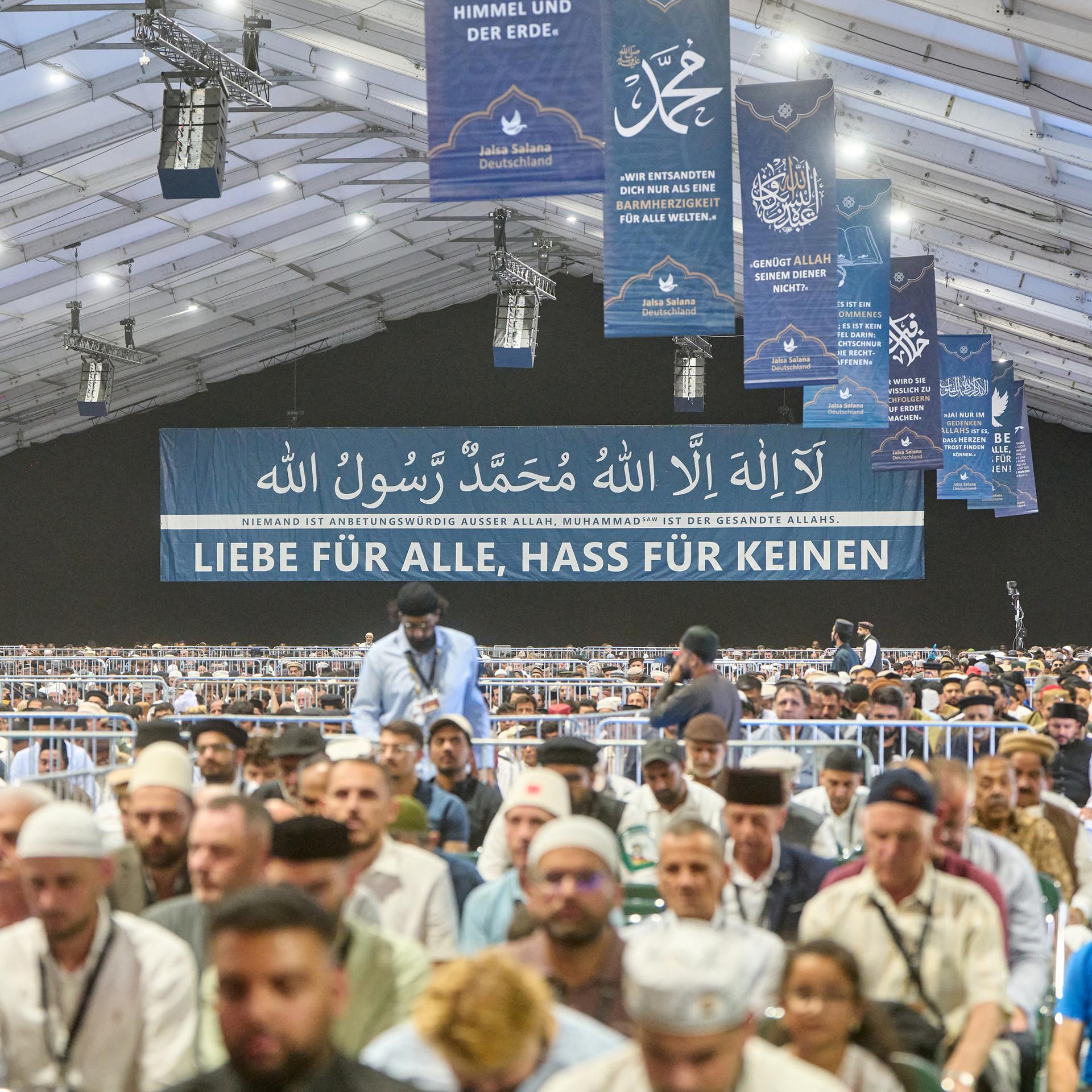 In einem großen Zelt sitzen muslimische Männer zum Gebet. Im Hintergrund ist ein großes Banner. Darauf steht: Liebe für alle, Hass für keinen.
