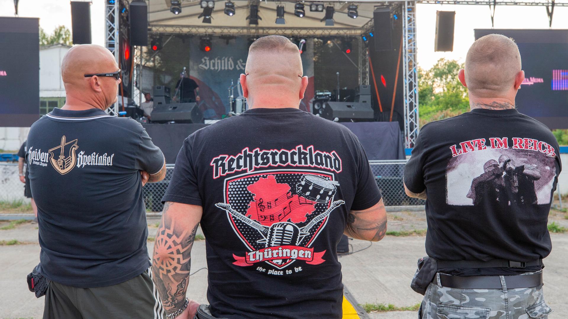 Drei Männer stehen auf dem Areal des "Schild und Schwert" Festival in T-Shirts mit den Aufdrucken Rechtsrockland (M) und "LIVE IM REICH" (r). Auf dem Festival treten rechtsextreme Bands auf.