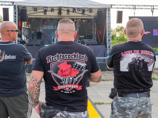 Drei Männer stehen auf dem Areal des "Schild und Schwert" Festival in T-Shirts mit den Aufdrucken Rechtsrockland (M) und "LIVE IM REICH" (r). Auf dem Festival treten rechtsextreme Bands auf.