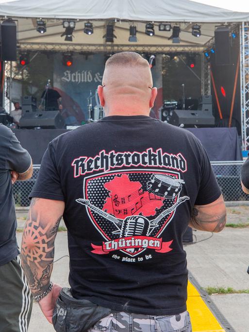 Drei Männer stehen auf dem Areal des "Schild und Schwert" Festival in T-Shirts mit den Aufdrucken Rechtsrockland (M) und "LIVE IM REICH" (r). Auf dem Festival treten rechtsextreme Bands auf.