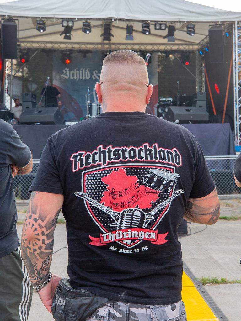 Drei Männer stehen auf dem Areal des "Schild und Schwert" Festival in T-Shirts mit den Aufdrucken Rechtsrockland (M) und "LIVE IM REICH" (r). Auf dem Festival treten rechtsextreme Bands auf.