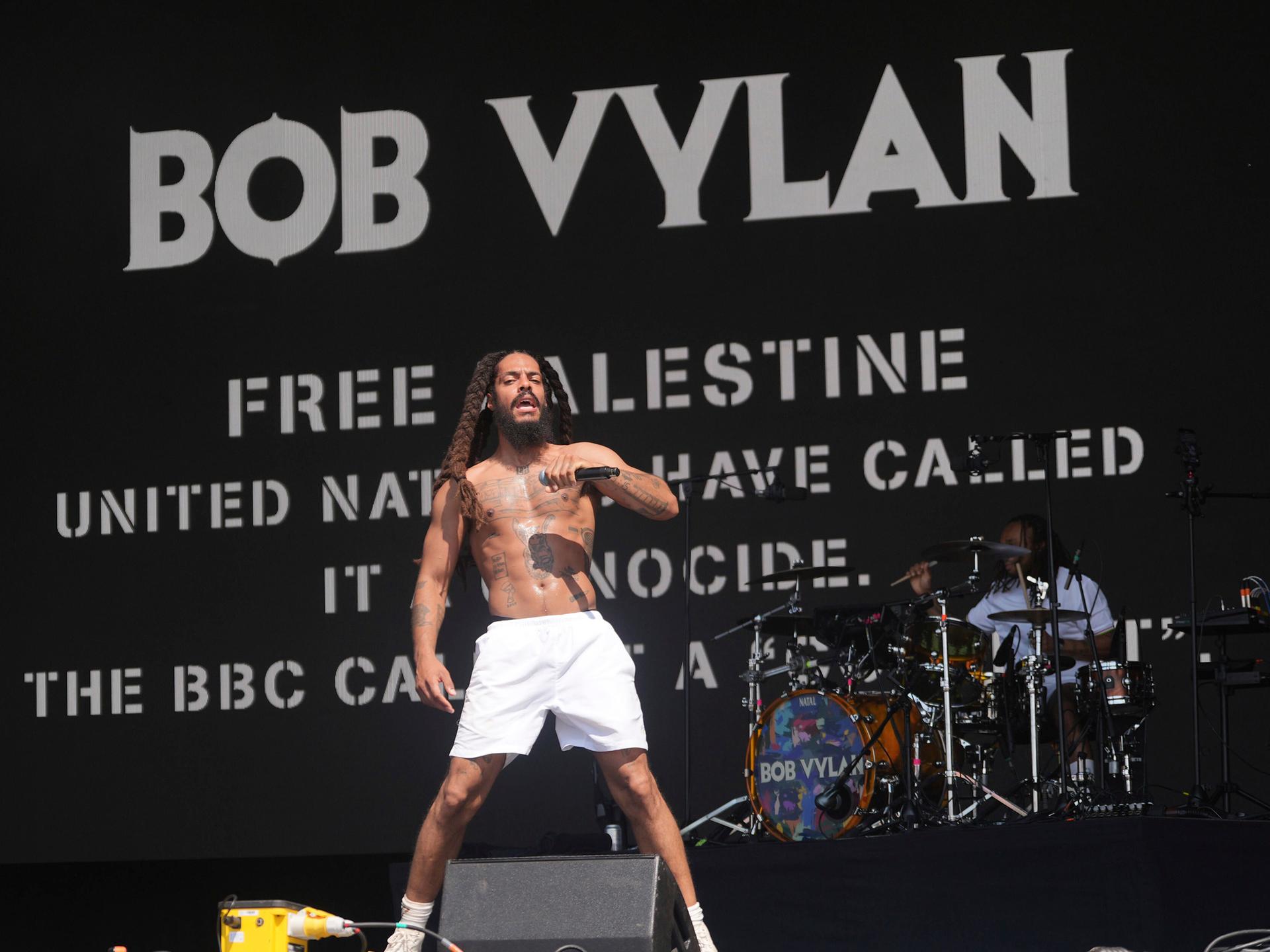 Vylan steht mit nacktem Oberkörper und nur mit weißen Shorts bekleidet auf der Bühne und singt in ein Mikrofon. Dahinter sein Schlagzeuger. Auf einer schwarzen Videowand dahinter stehen in weißer Schrift "Free Palestine" und andere pro-palästinensische Slogans.
