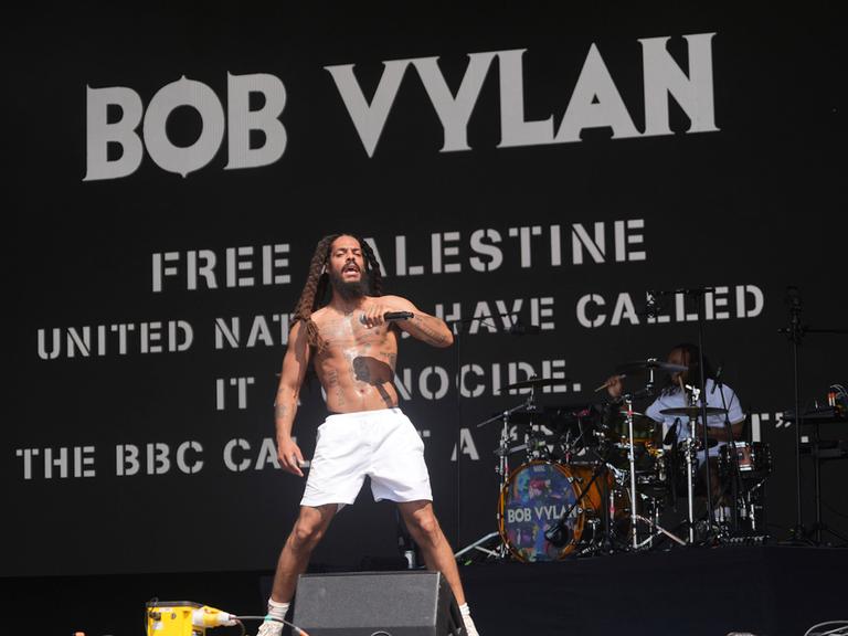 Vylan steht mit nacktem Oberkörper und nur mit weißen Shorts bekleidet auf der Bühne und singt in ein Mikrofon. Dahinter sein Schlagzeuger. Auf einer schwarzen Videowand dahinter stehen in weißer Schrift "Free Palestine" und andere pro-palästinensische Slogans.