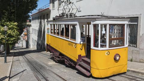 Eine gelbe Straßenbahn fährt eine steile, gepflasterte Straße entlang vor hellen Gebäuden.